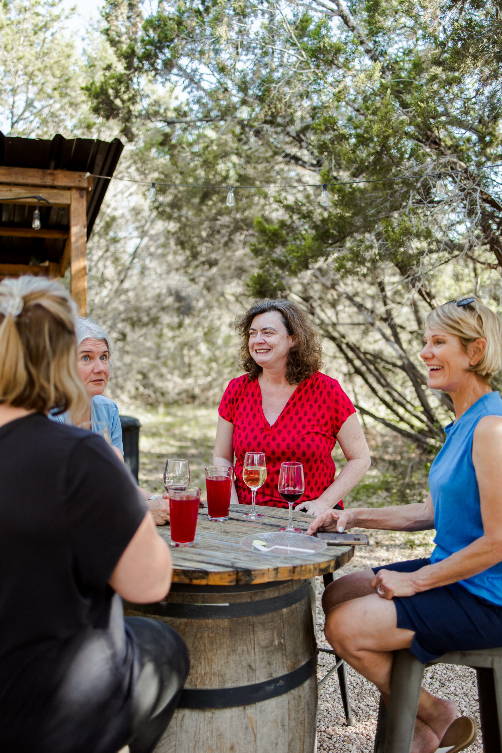 Four women sit around a rustic outdoor table made from a barrel, smiling and chatting with drinks in front of them, enjoying the sunlight and trees—an ideal afternoon at a Women's Ranch Retreat.