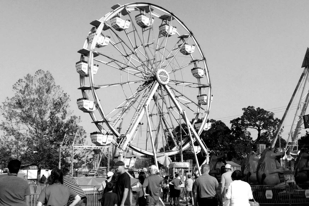 Black and white photo of people walking toward a large Ferris wheel at an outdoor fair, with trees and other amusement rides visible in the background.