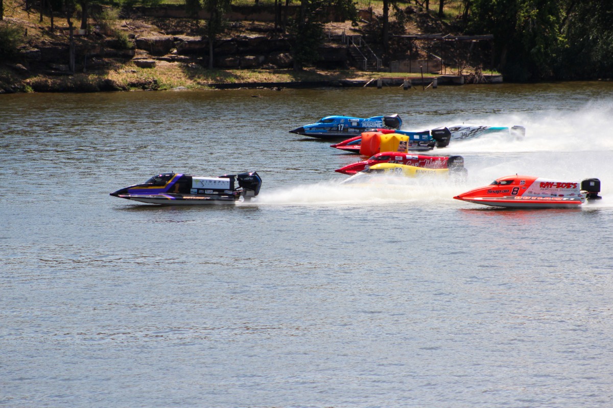Five colorful F1 powerboats race across Lake Marble Falls, creating white spray trails behind them. Trees and grassy banks are visible in the background under Colorado’s sunny sky.