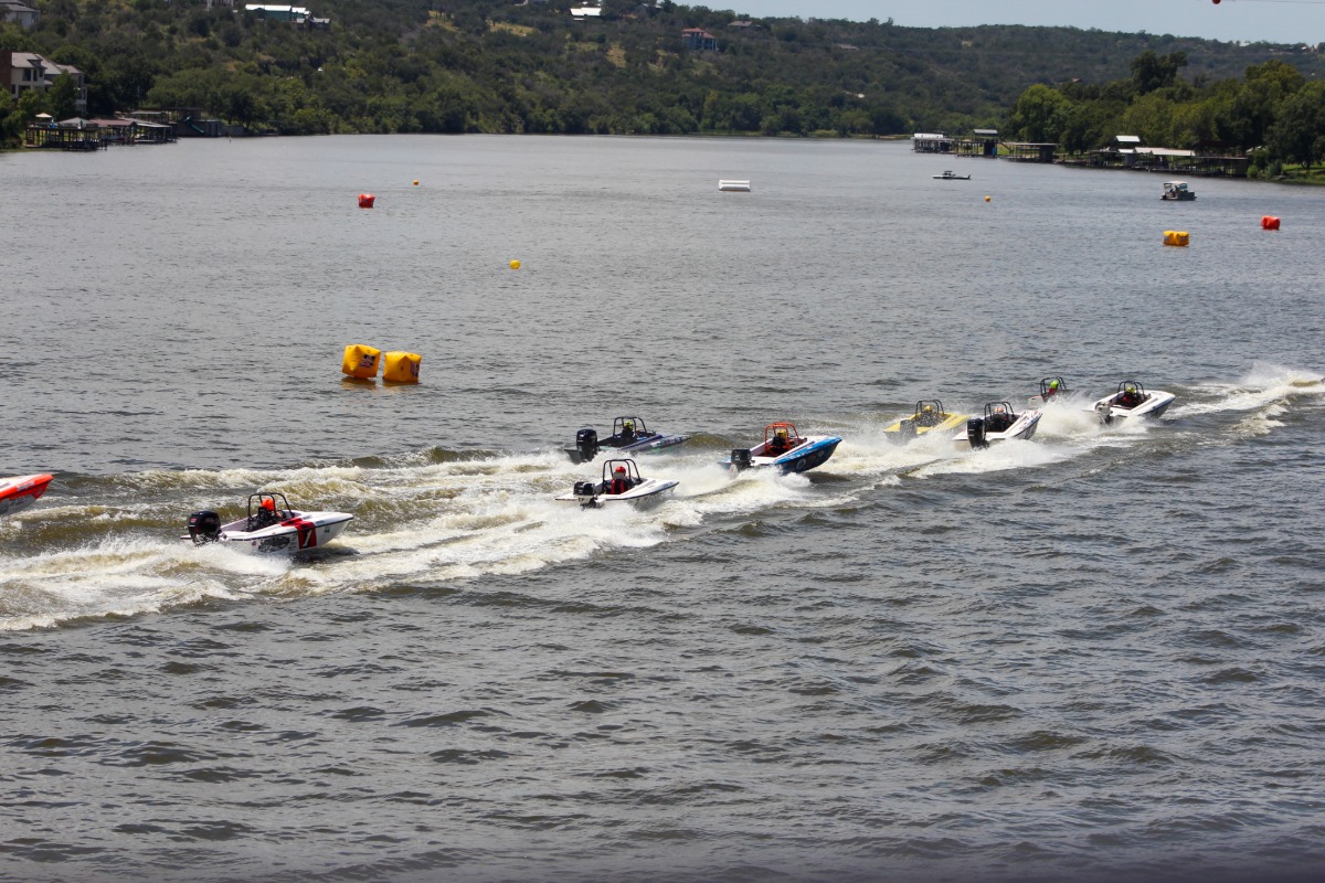 F1 powerboats with drivers race on Lake Marble Falls, creating white trails in the water. Yellow buoys mark the course, while green hills dotted with houses form a scenic backdrop.