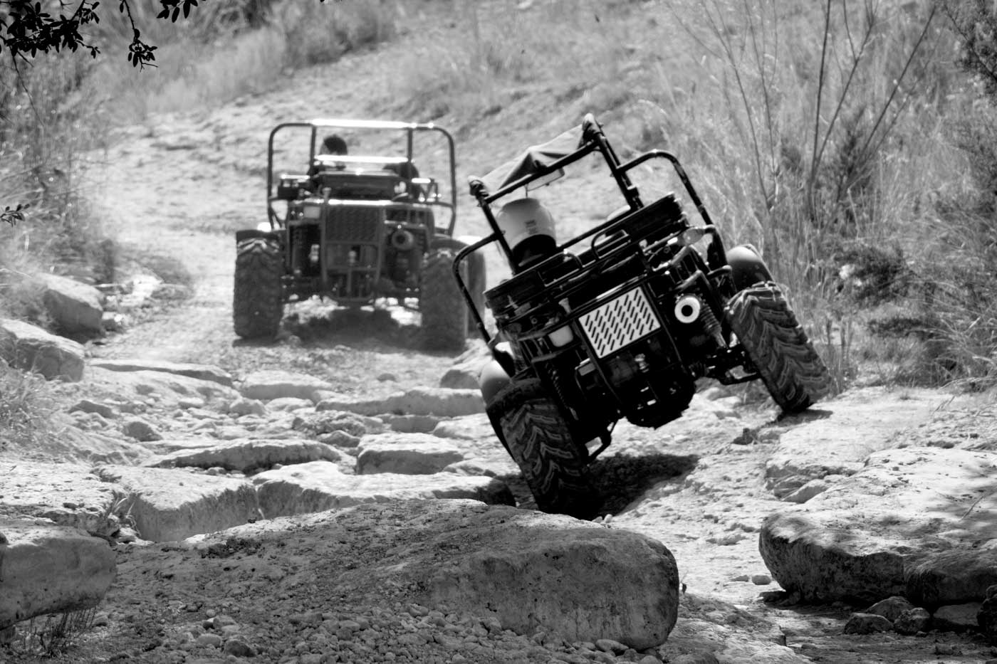 Two off-road vehicles navigate a rocky, uneven trail in Burnet County, the lead one tilted as it tackles rough terrain. This black and white image captures the adventurous spirit of a perfect Guys Weekend or 3 Day Itinerary outdoors.