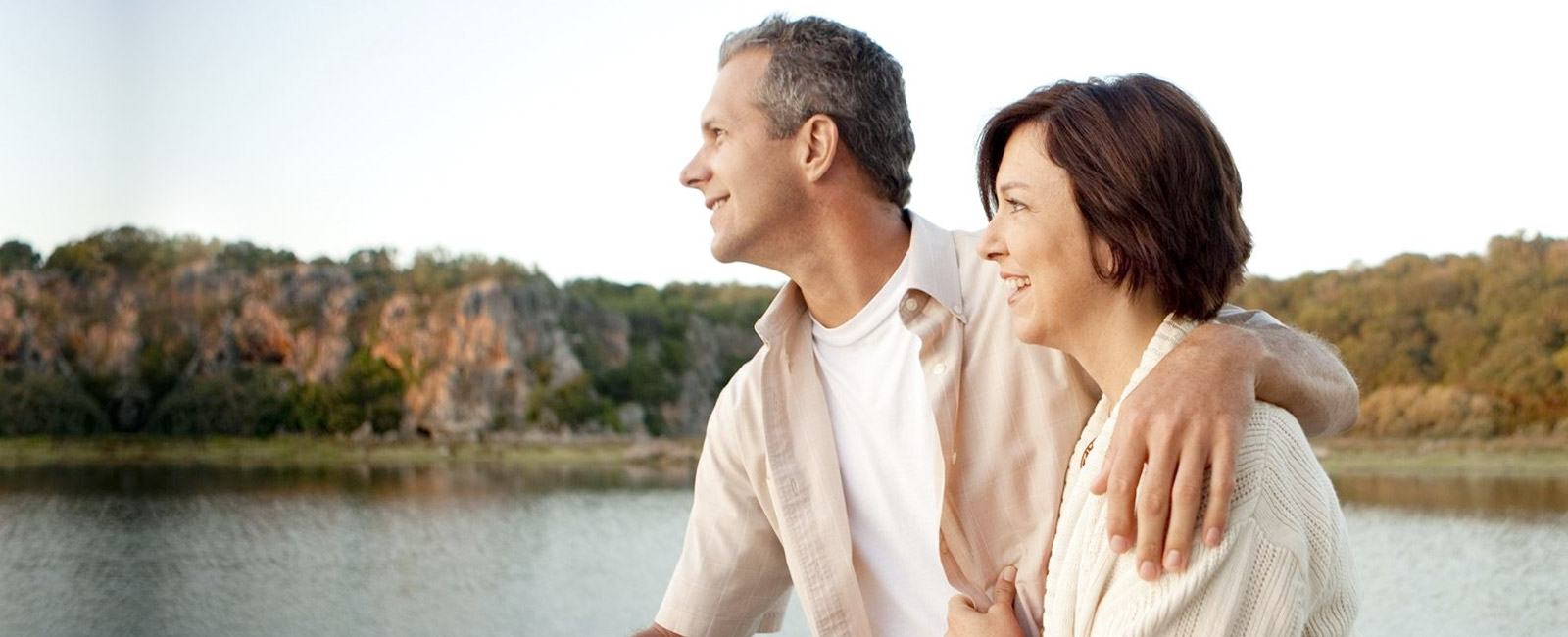 A middle-aged couple stands by a calm lake during a test event, smiling and looking into the distance. The man has his arm around the woman, with trees and rocky hills in the background.