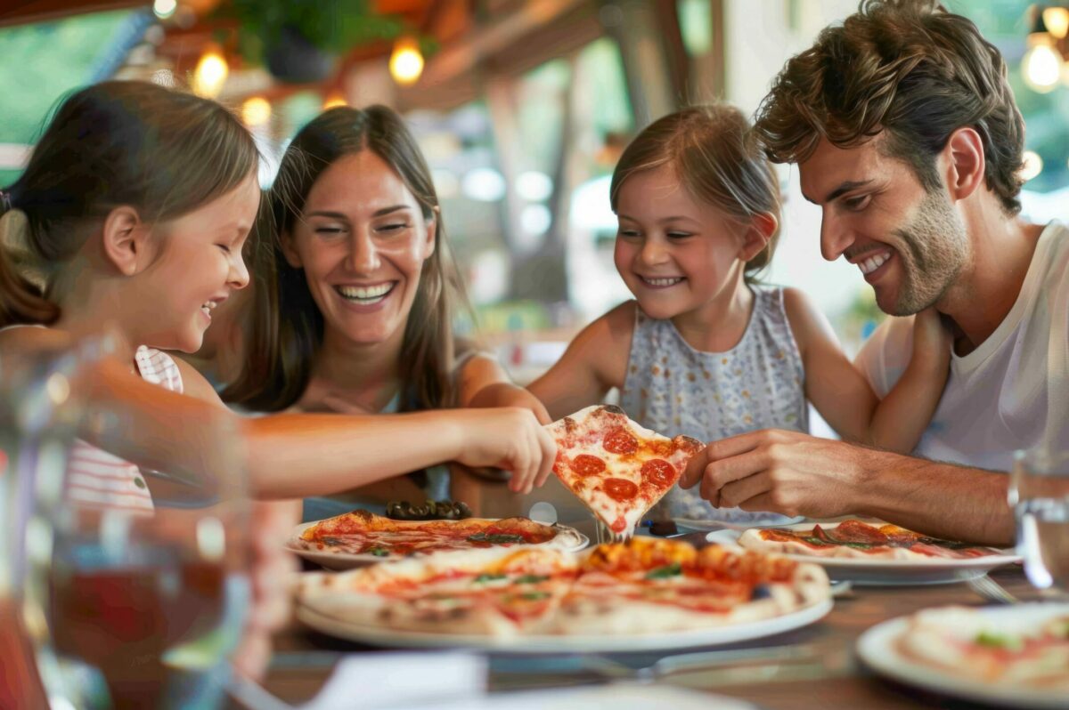 A happy family of four, including two young girls and two adults, sits at a restaurant table enjoying and sharing slices of pizza, smiling and laughing together. Glasses and plates are on the table in a cozy, warmly lit setting.