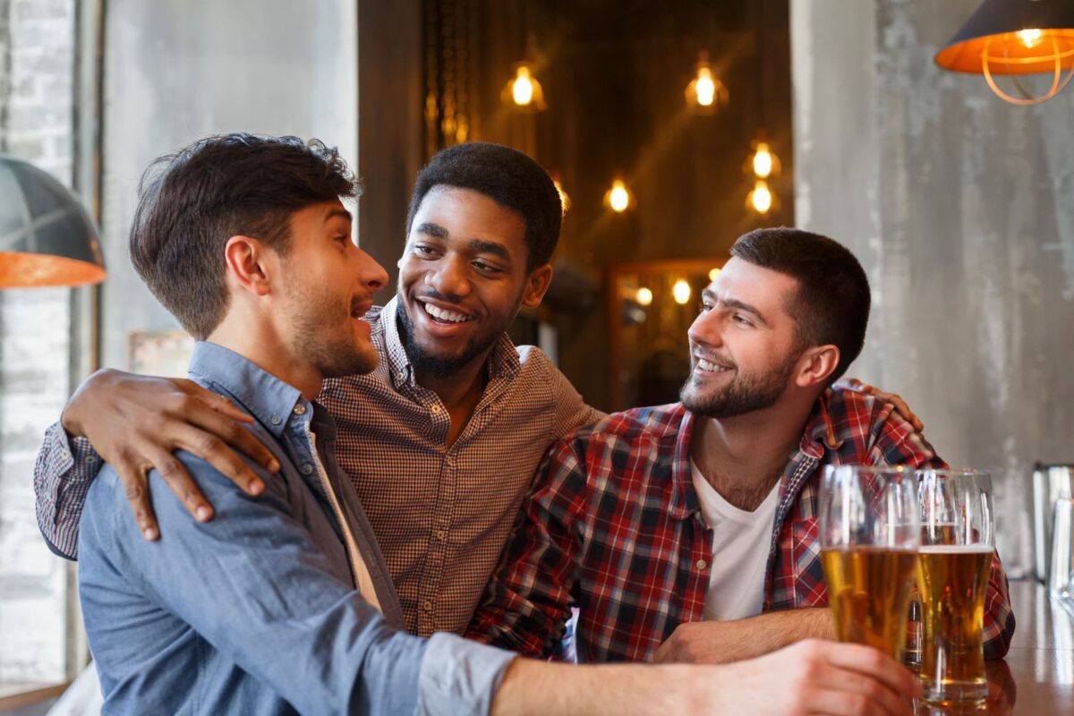 Three young men sit at a bar, smiling and laughing together. One has his arm around another, and all have glasses of beer in front of them. Warm lighting and hanging bulbs create a cozy, inviting atmosphere.