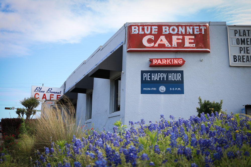 A white building with red signs reading Blue Bonnet Cafe and Pie Happy Hour, surrounded by blooming bluebonnet flowers under a partly cloudy sky—an ideal stop for your Guys Weekend in Burnet County.