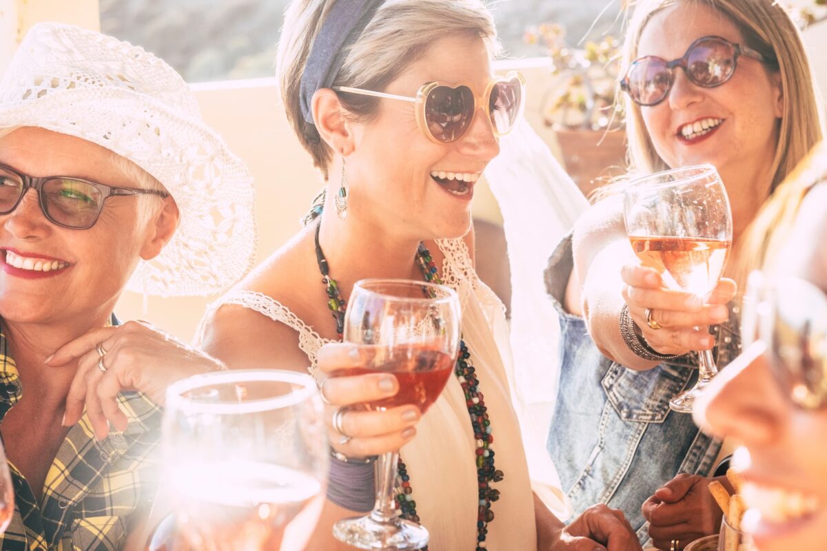 A group of women wearing sunglasses smile and toast with glasses of rosé wine, enjoying a sunny outdoor gathering.