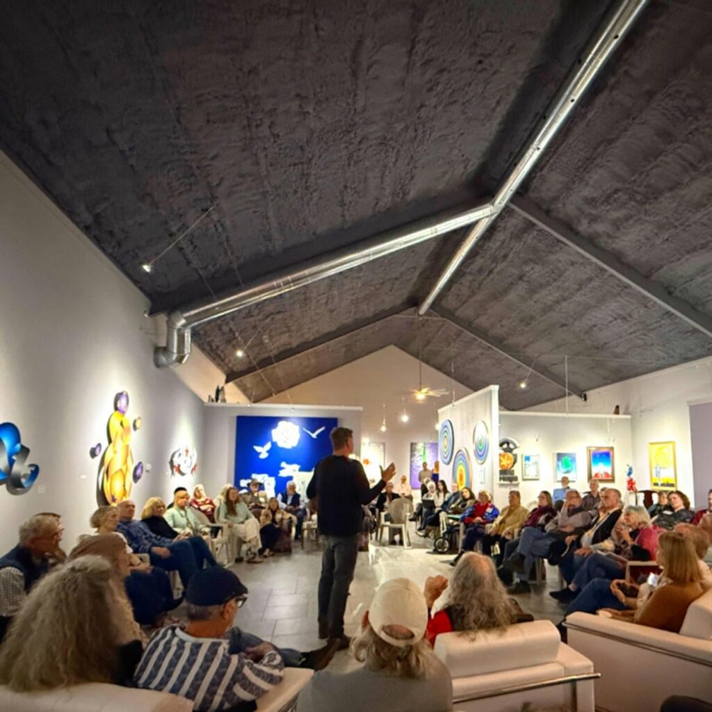 A man stands speaking to a seated audience in Museo Benini, an art gallery with colorful abstract paintings on white walls, under a high, dark ceiling.