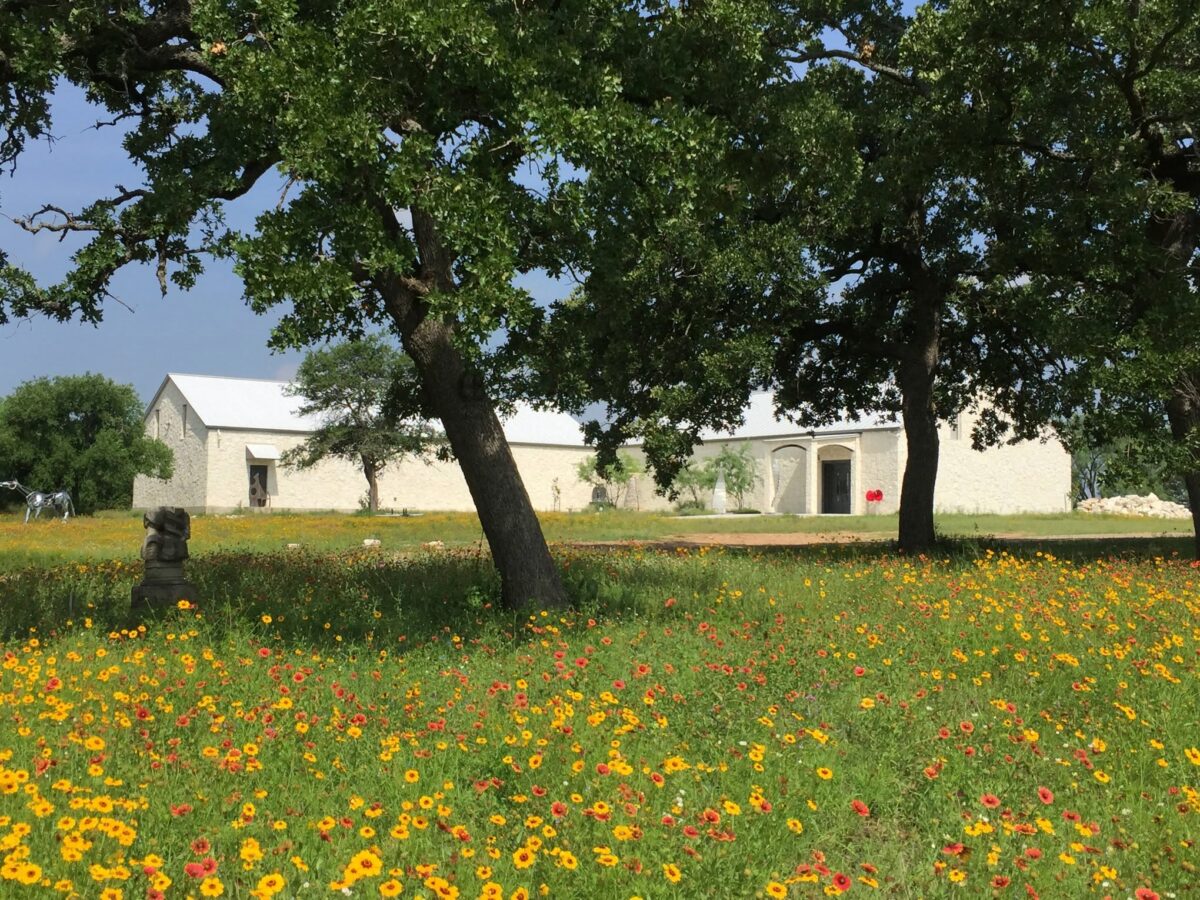 A stone building with a white roof sits behind large trees and a field of yellow and red wildflowers under a clear sky. A small stone statue, reminiscent of art found at the Museo Benini, is visible among the flowers in the foreground.